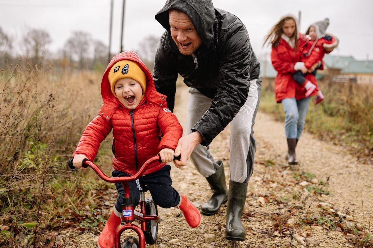 Bolgodan biciklizik a piros esőkabátos kisgyerek, akit az apukája segít